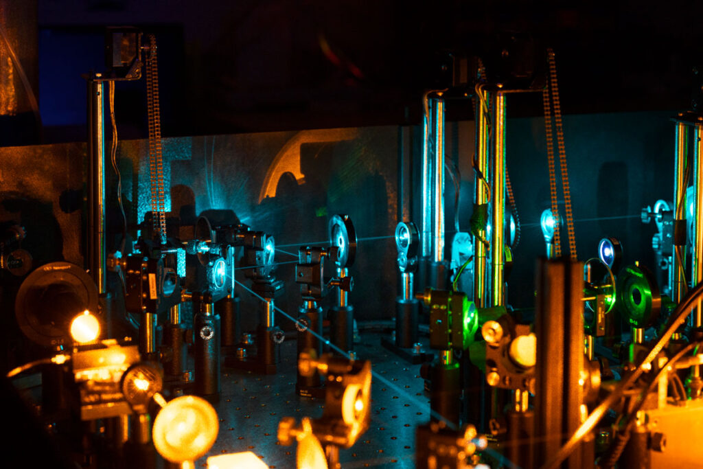 Close-up of optical equipment in a dark room illuminated by multicolored laser light. A clear blue laser beam travels from the front of the photo to the back right, refracting off of tiny lenses.