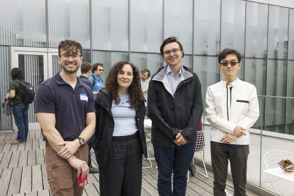 Four students stand together on an outdoor deck in front of the tall glass walls of the Singh Center. They are all smiling warmly and looking at the camera while other symposium guests mill about behind them.