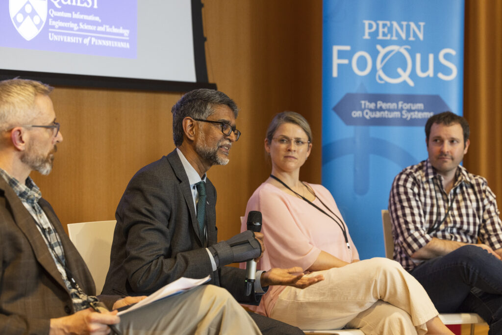 Four people sit in chairs in front of a blue Penn FoQuS banner. The second person from the left (Vijay Kumar) holds a microphone and speaks while gesturing with his other hand palm up. The other three people are looking at Vijay and listening with thoughtful expressions.